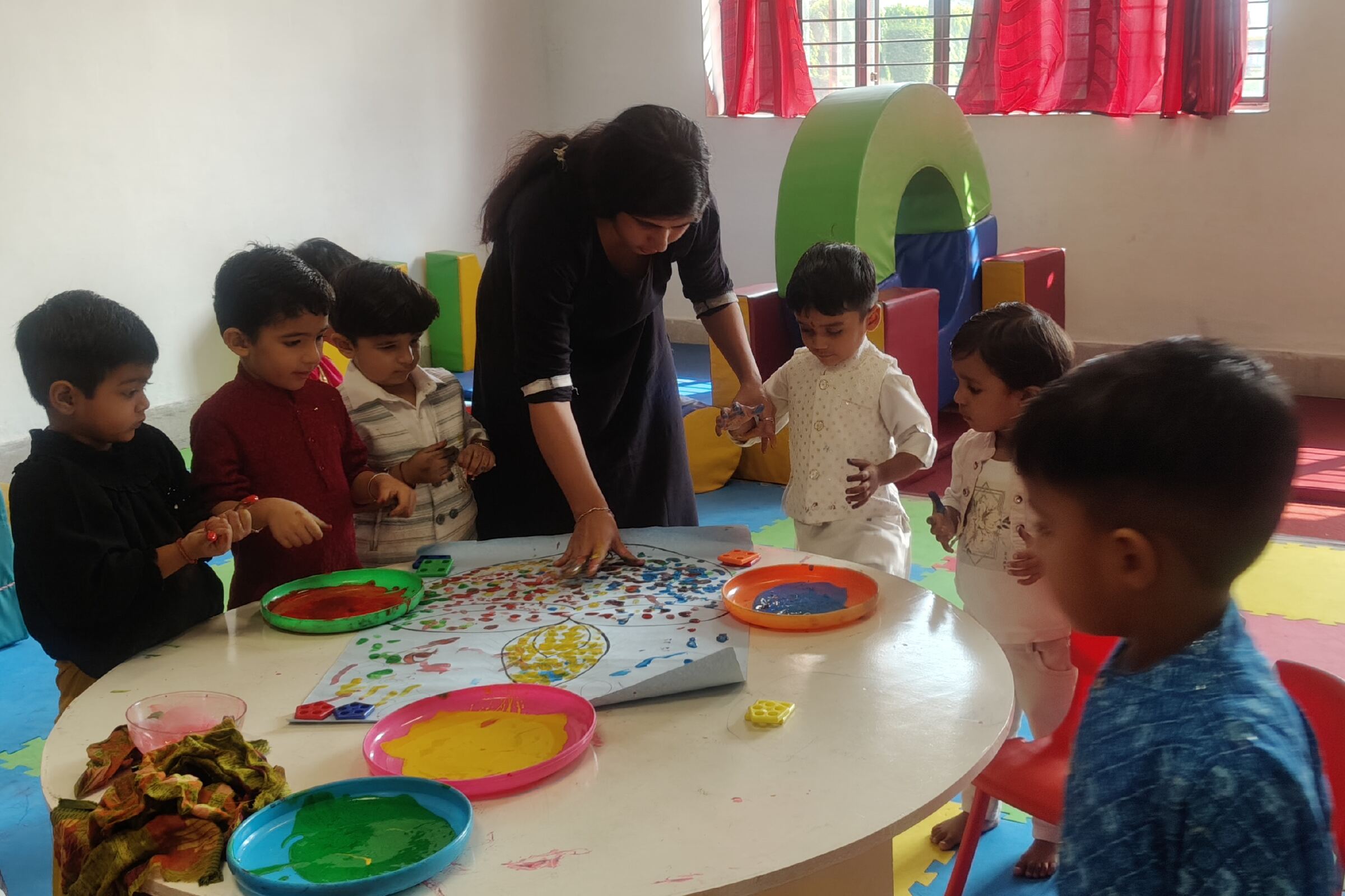 Children around a round table painting with their hands, a facilitator among them, bowls of colour on the table.
