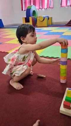 A small child crouching on a red carpet, reaching to place a piece on top of a tall tower of coloured wooden rings.