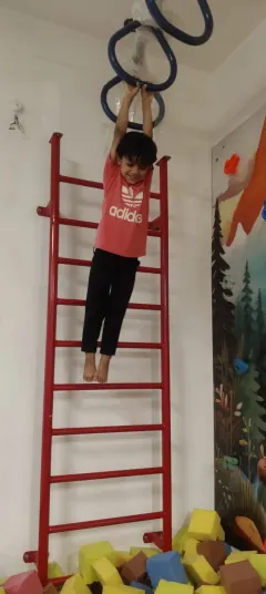 A child hanging by both hands from rings near the ceiling, above a pit of foam blocks.