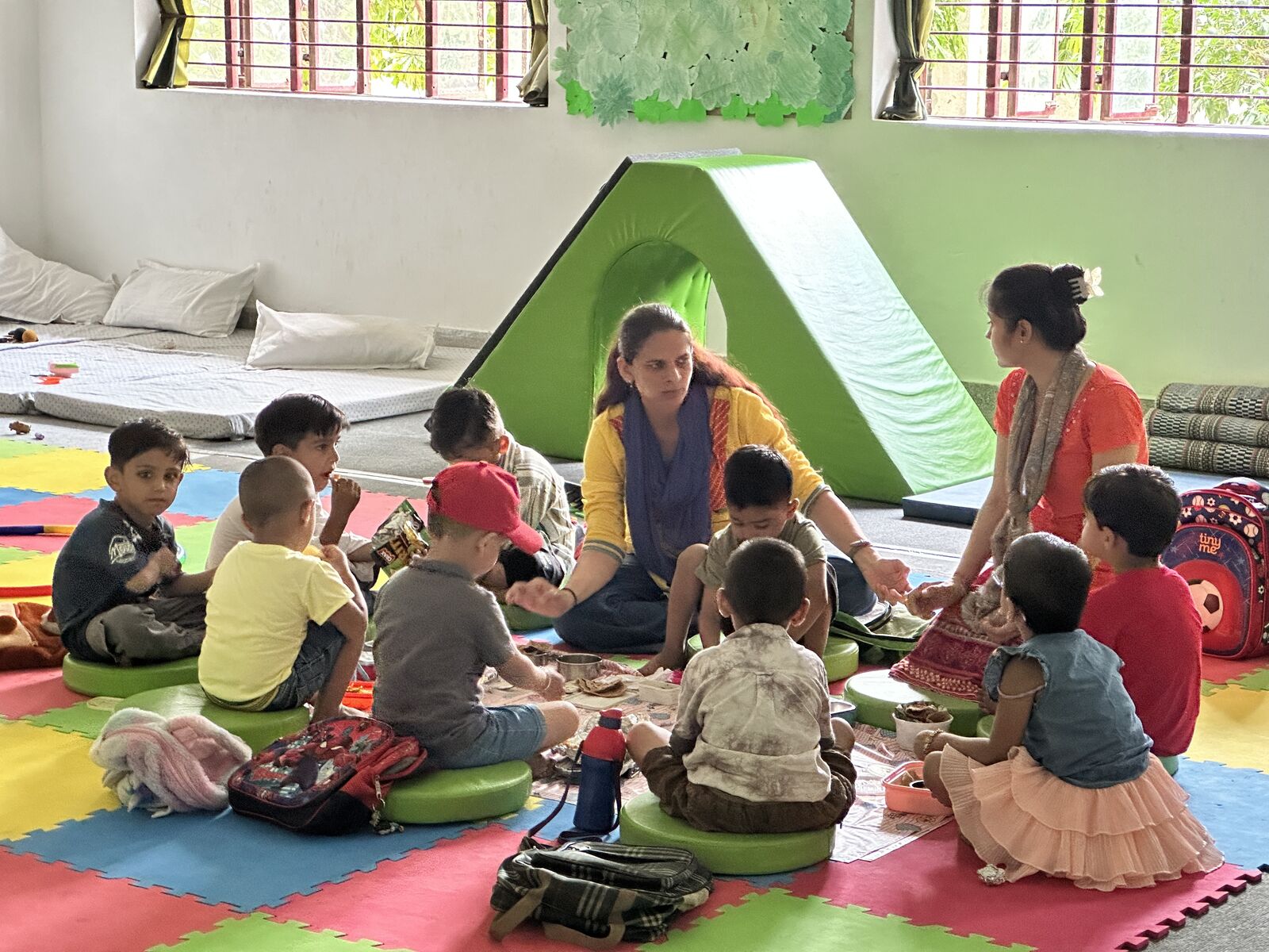 Children eating lunch in a circle on the classroom floor, two facilitators sitting with them.