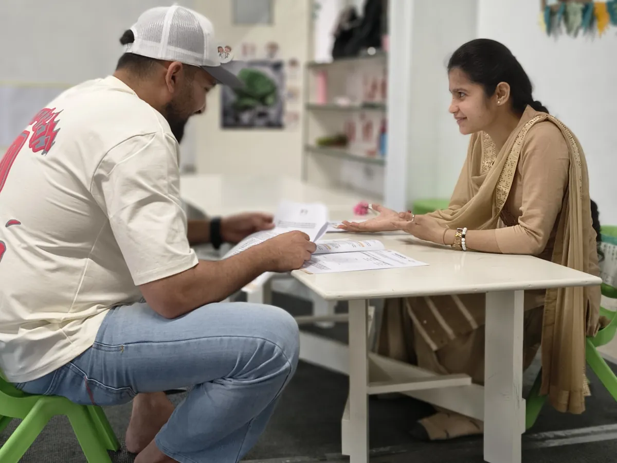 A father in a white cap reads a printed report at a low classroom table, while a facilitator sitting across from him smiles and talks him through it.