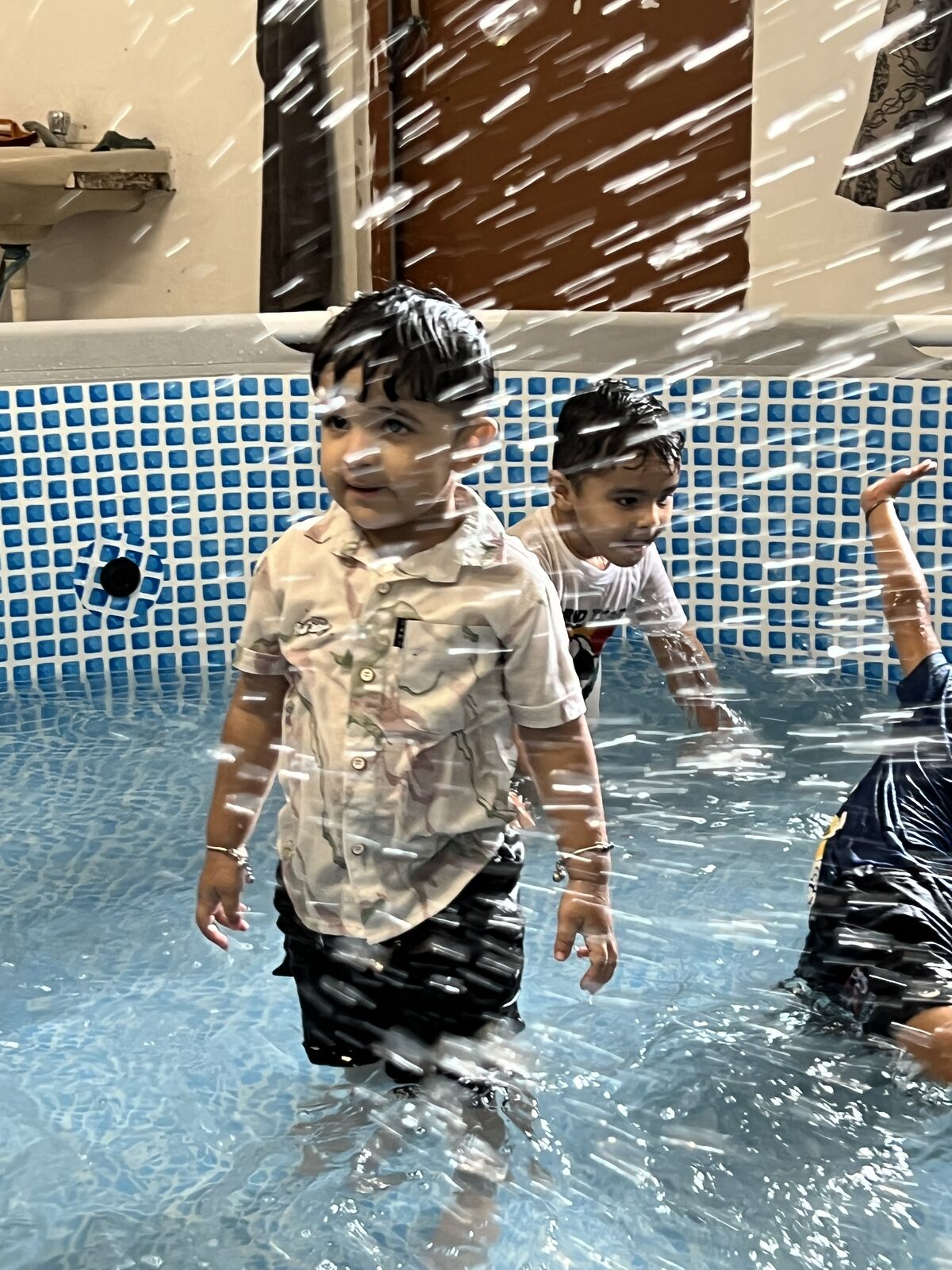 Two children standing in shallow water in a small tiled pool while water splashes all around them.