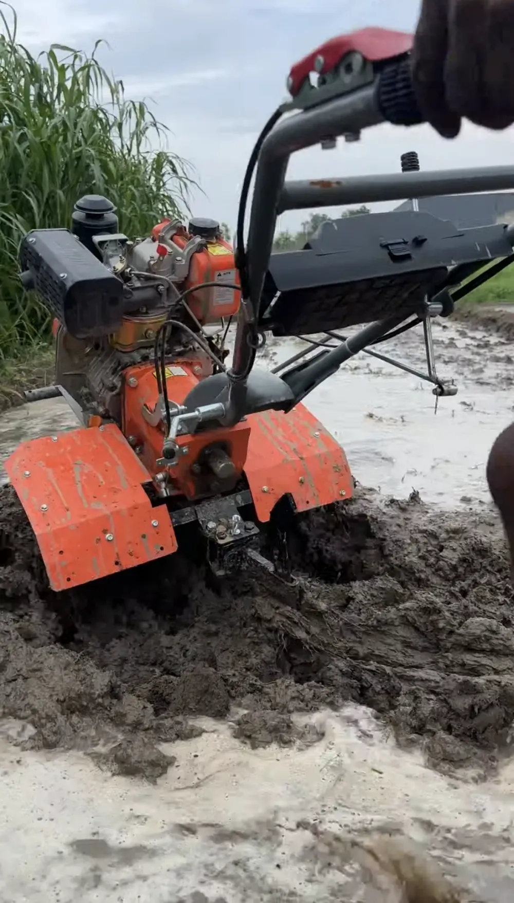 An orange power tiller churning through a flooded, muddy field at Samsara, a hand on its handlebar and tall grass behind.