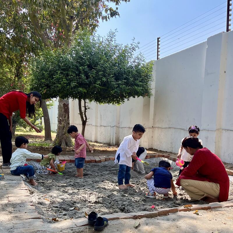 Children digging in the sandpit under the trees, two facilitators nearby.