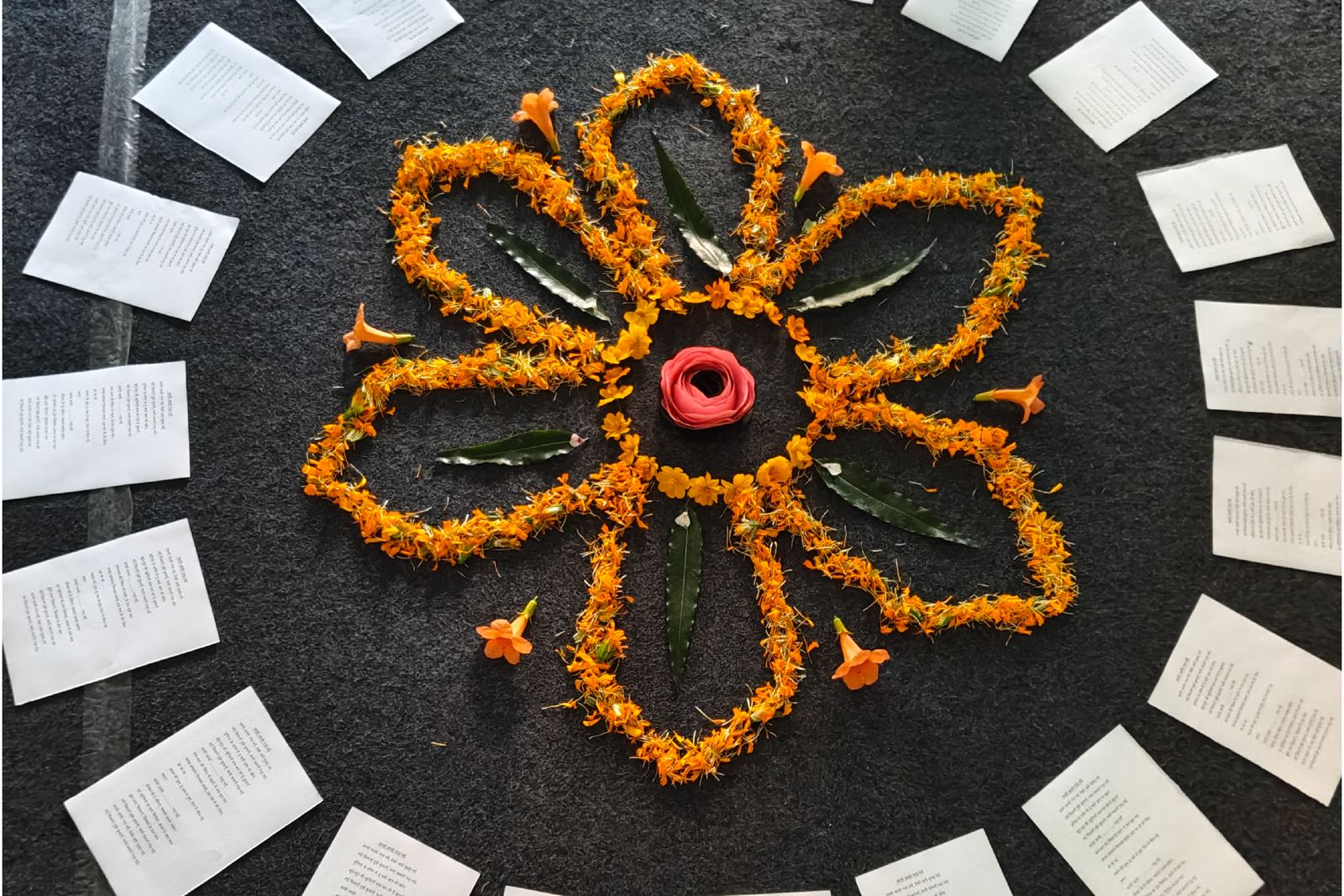 A flower made of marigold petals and leaves on a dark cloth, ringed by printed pages.