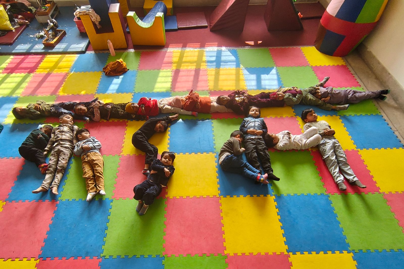 Children lying head to toe in a long line across the coloured mat, making a shape with their bodies.