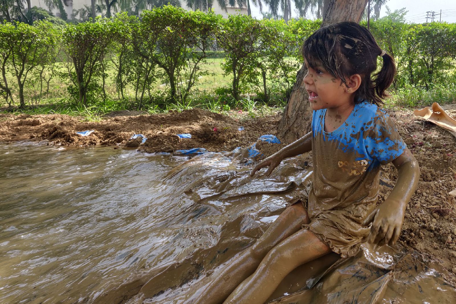 A child sitting in muddy water at the edge of the field, laughing.