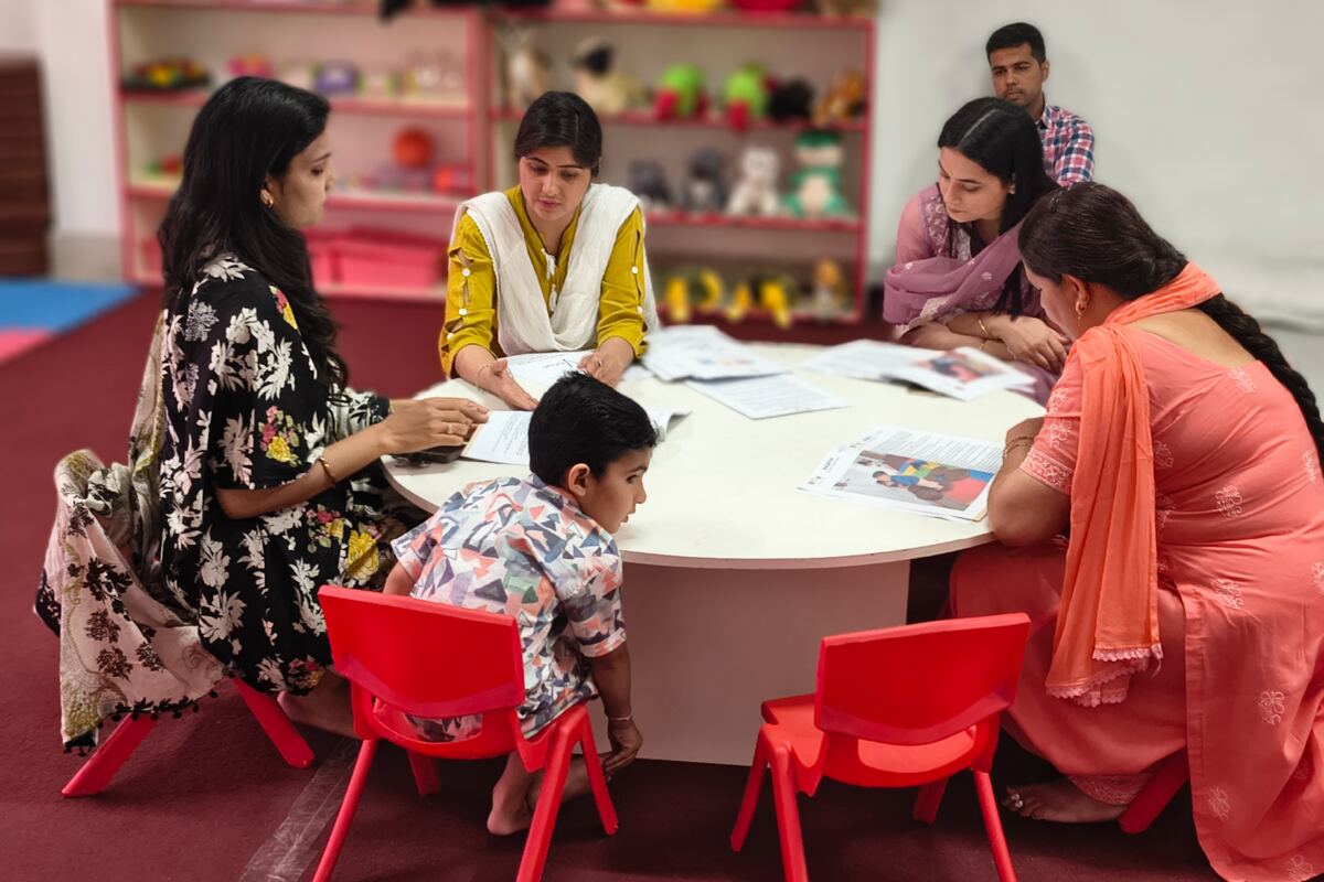 Facilitators and parents around a round table in the classroom, a child on a red chair among them.