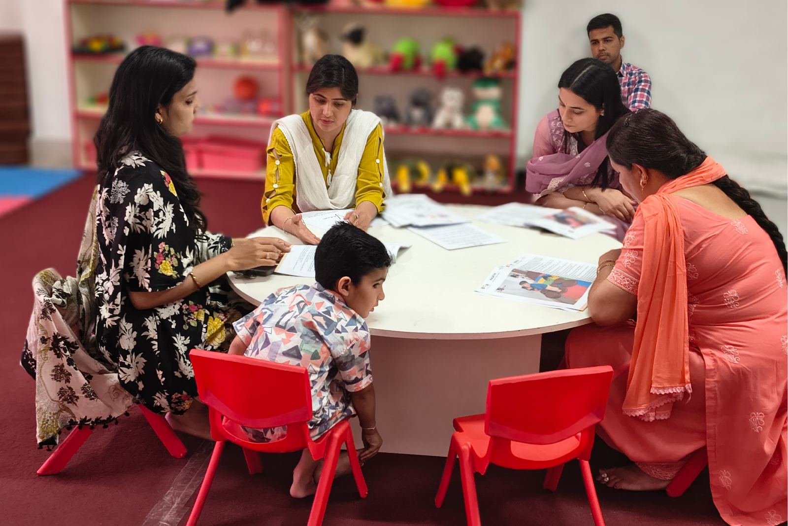 Facilitators and parents around a round table, a child on a red chair among them.