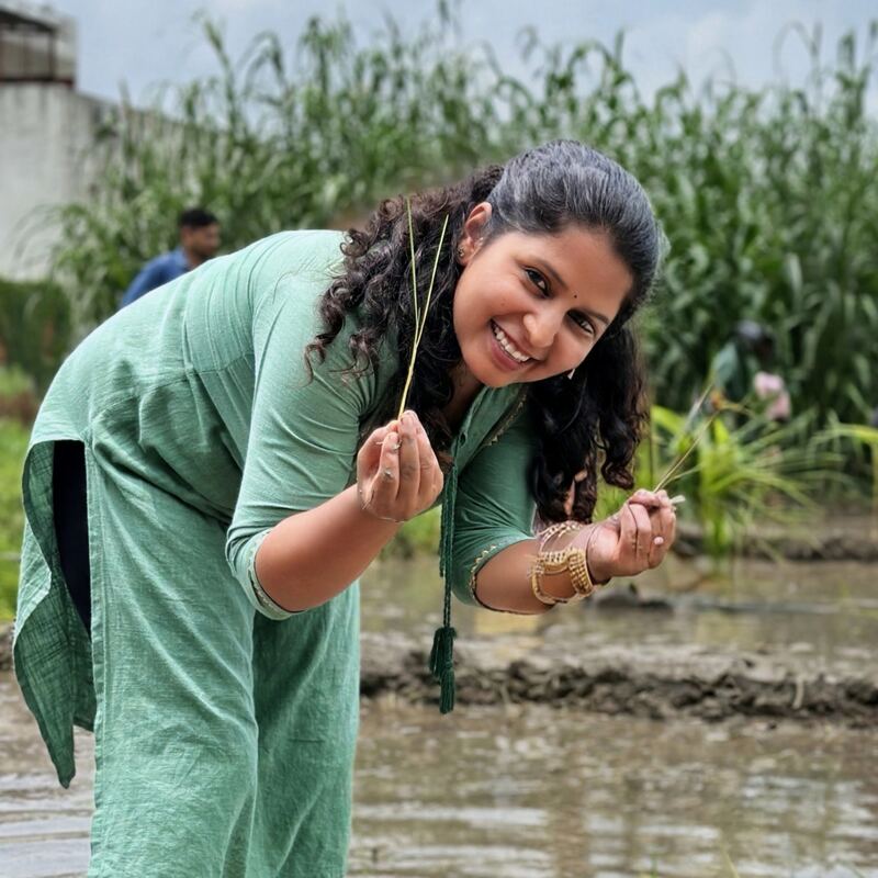 Sunita, bending over the flooded paddy field with a rice seedling in her hand, smiling.
