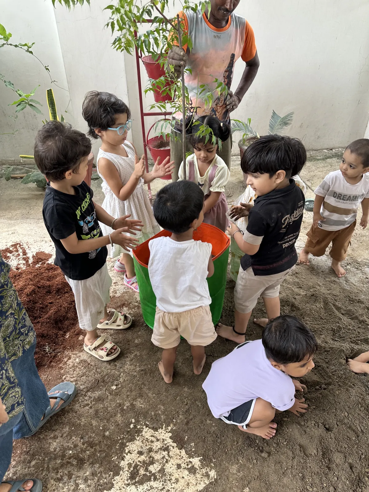 Children crowd round a green grow bag, hands in the soil, as a gardener holds a young tree upright for them to plant.